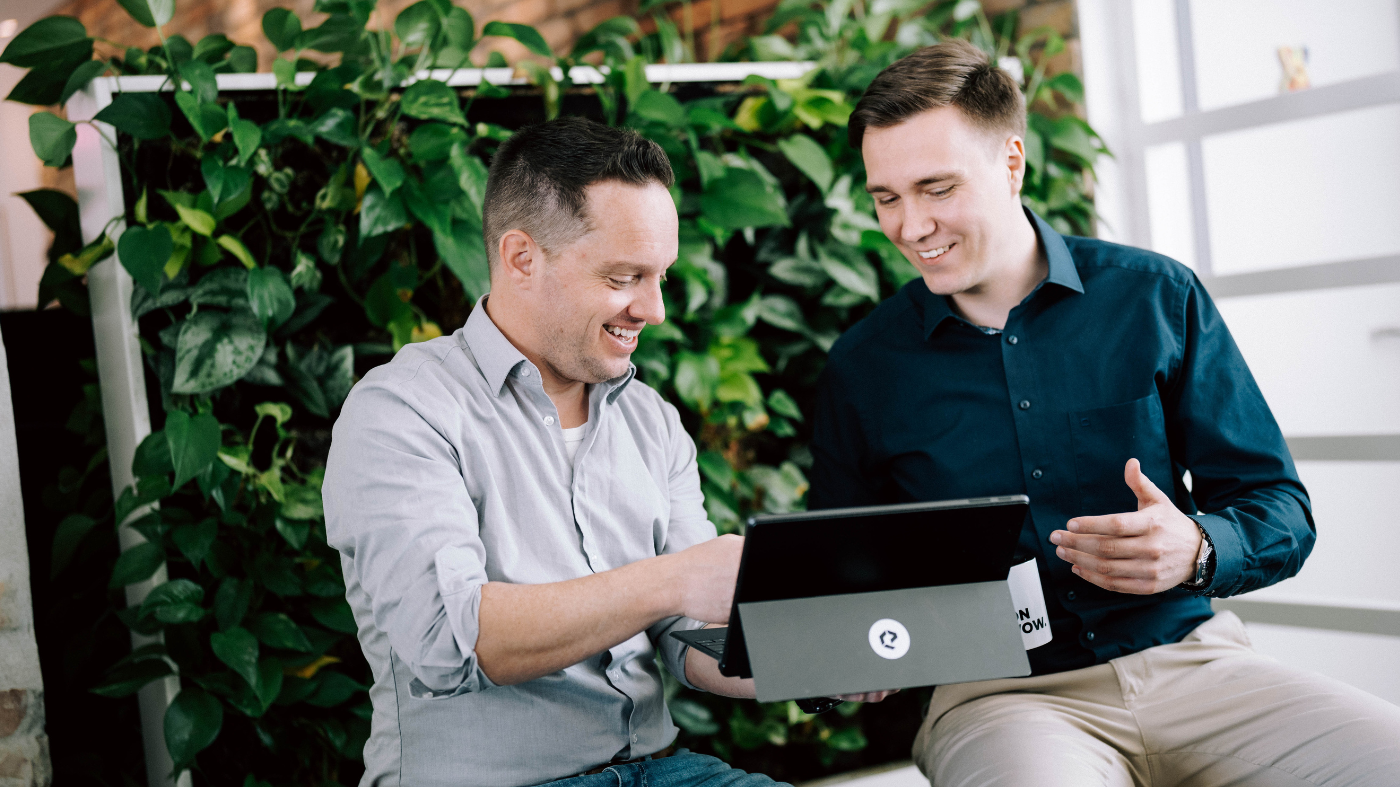 Relaunchagentur aus Süddeutschland Zwei Männer sitzen vor einer grünen Pflanzenwand und schauen lächelnd auf ein Tablet.